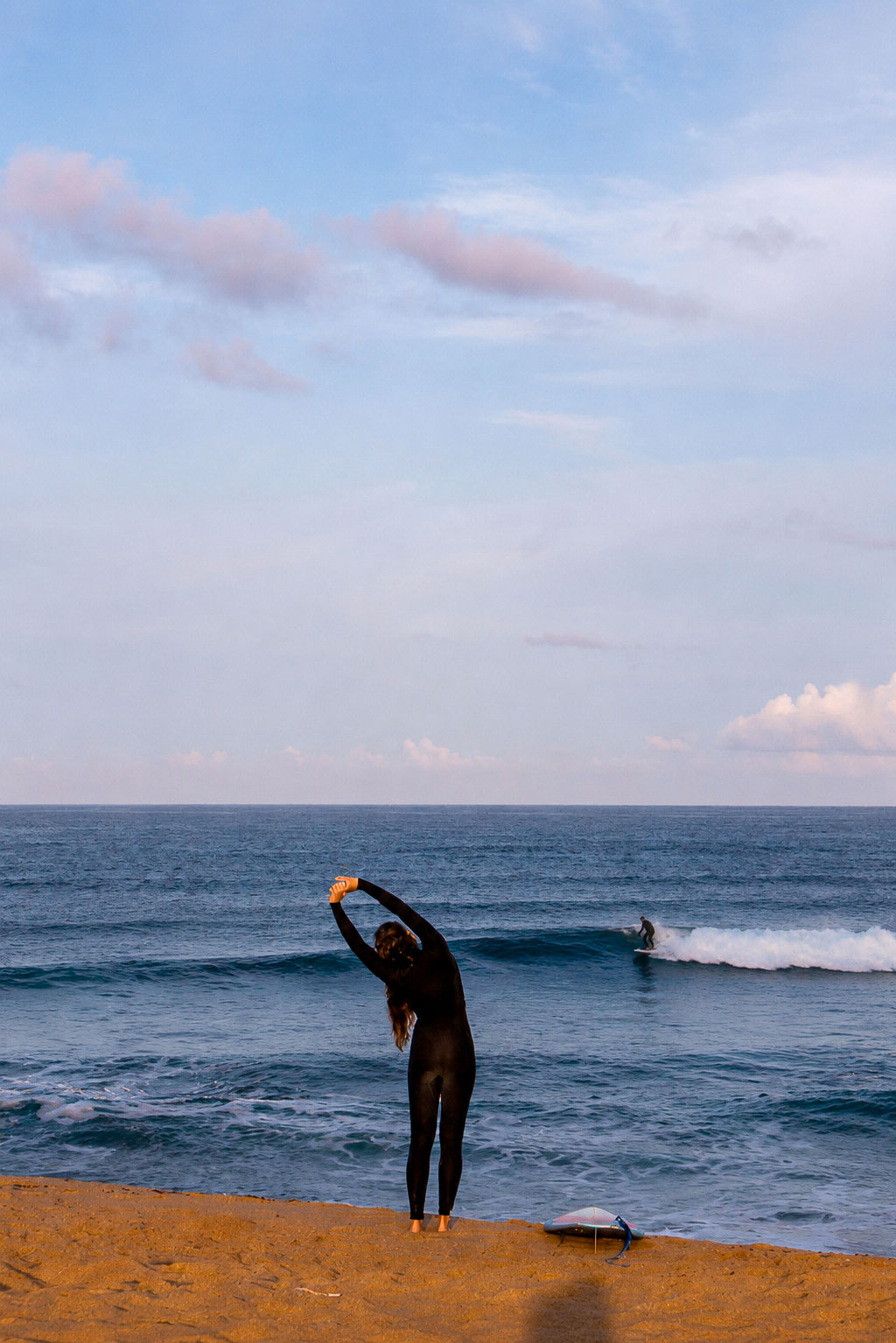 Surfer stretching on the beach before a surf session in Ericeira, Portugal, while another surfer rides a small wave at golden hour.